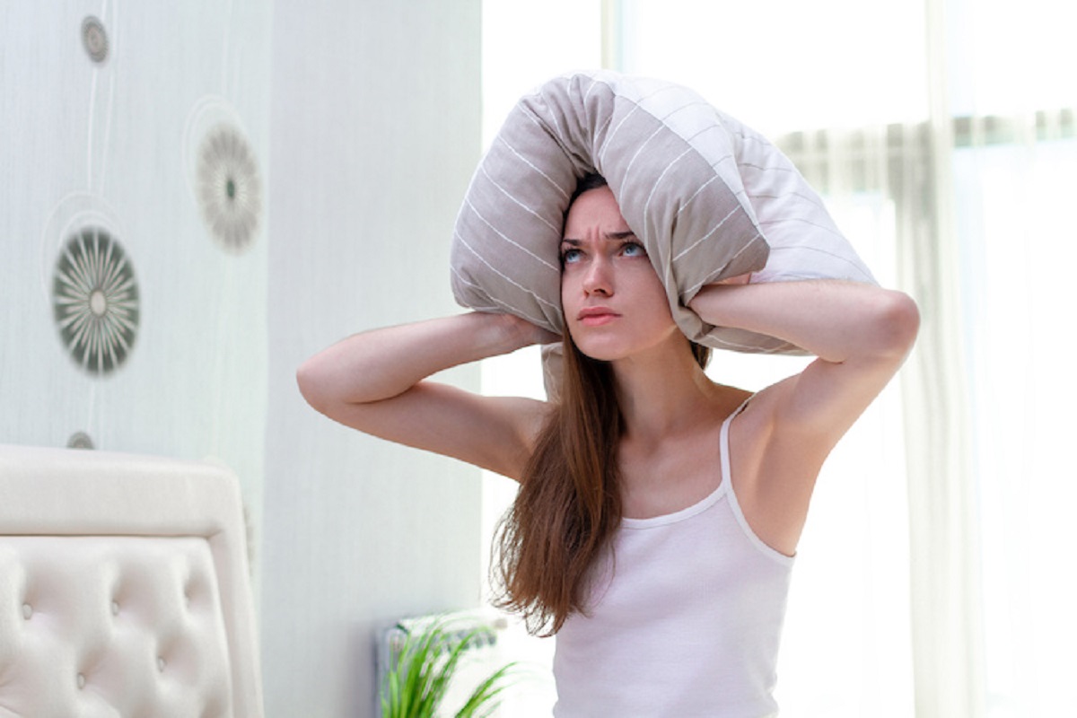 Angry woman suffering and disturbed by noisy neighbors and covering her ears with pillow while trying to falling sleep in bed at home in early morning